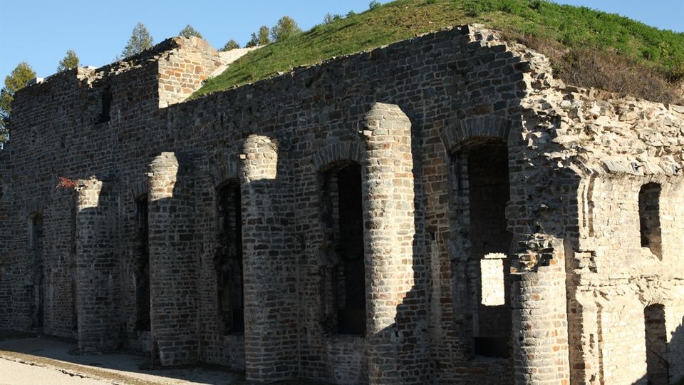 Rovine di una struttura fortificata in pietra con pilastri e archi murati lungo la facciata e aperture rettangolari. Sullo sfondo si vede un terrapieno erboso sopra le mura e un cielo sereno.