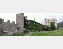 Cortile di un complesso storico con mura e torri in pietra, affiancato da un edificio basso ricoperto di edera con finestre e balconi. Sullo sfondo si vede un grande edificio in pietra chiara tipo castello e un prato verde ai piedi di colline boscose.