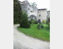 Edificio storico in pietra e intonaco, simile a un piccolo castello o fortificazione, con alte mura e un grande portale ad arco. Davanti si estende un prato con cespugli e alberi, attraversato da un sentiero sterrato curvo.
