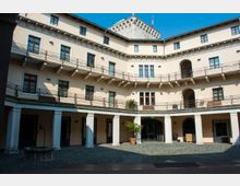 Cortile interno di un edificio storico a pianta semicircolare, con facciate intonacate chiare, finestre regolari e ballatoi con ringhiere metalliche su più livelli. Al piano terra portico con colonne, pavimentazione in ciottoli e un pozzo centrale, con una torre in pietra che sporge sullo sfondo sotto cielo azzurro.