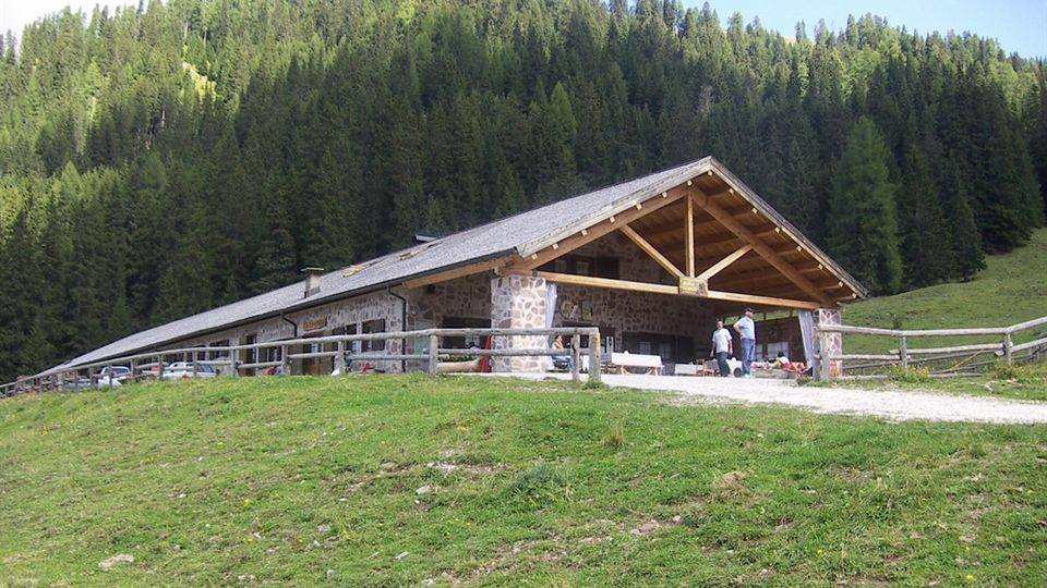 Rifugio di montagna in pietra e legno con ampio portico e tetto spiovente, affacciato su un prato recintato. Sullo sfondo si vede un fitto bosco di conifere che copre il versante della montagna.