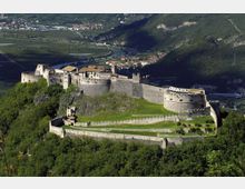 Veduta panoramica di un grande castello medievale in pietra su una collina, con mura merlate, torri e bastioni che circondano un ampio prato interno. Sullo sfondo si vede una valle con fiume sinuoso, campi coltivati, strade e un piccolo centro abitato ai piedi di montagne boscose.