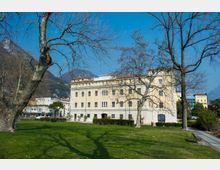 Edificio storico a più piani con facciata chiara e finestre regolari, affacciato su un ampio prato di parco con alberi spogli e un vialetto con panchina. Sullo sfondo si vedono montagne e altri edifici sotto un cielo sereno.