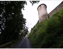 Strada asfaltata in salita tra alberi e vegetazione, che costeggia un alto muro in pietra. In alto si vede una torre cilindrica di una fortificazione, con sommità scura e piccole finestre.