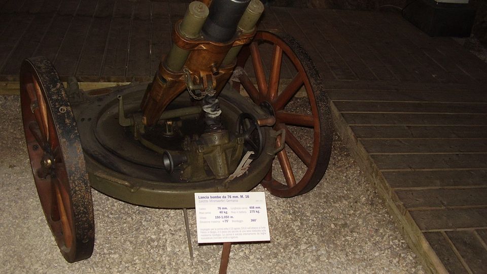 Interno di un museo con pavimento in ghiaia e passerella in legno, dove è esposto un pezzo di artiglieria su ruote con un cartello informativo davanti. Sullo sfondo si vede una parete scura in pietra e un’area illuminata in modo soffuso.