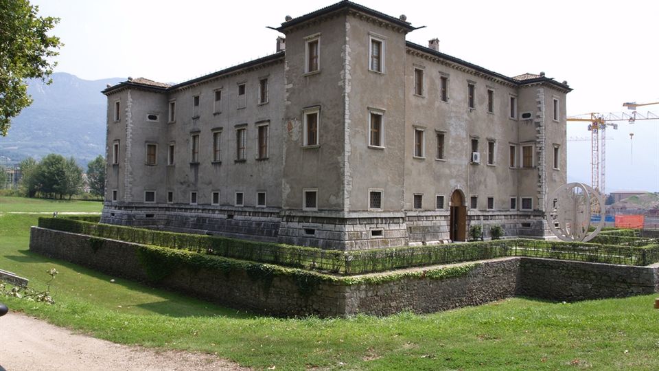 Edificio storico in muratura a pianta quadrata, con tetto a padiglione e numerose finestre, circondato da un fossato e da bassi muretti con siepi. La struttura si trova in un’area verde pianeggiante con montagne sullo sfondo.