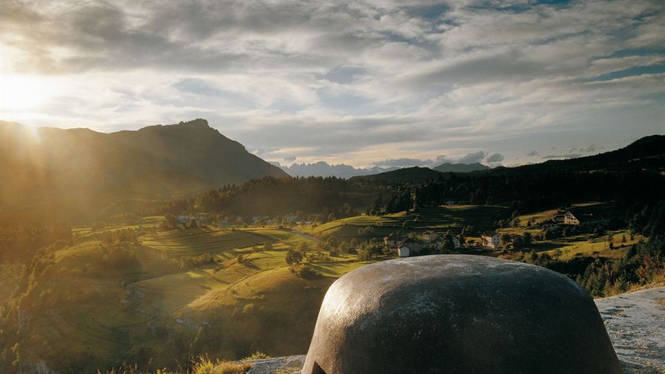 Veduta panoramica su una valle montana con prati e piccoli borghi, illuminata dal sole basso, con catene montuose sullo sfondo e cielo molto nuvoloso. In primo piano, su una sporgenza rocciosa, si vede una piccola struttura a cupola in cemento con feritoie, simile a un bunker.