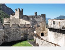 Cortile interno di un castello medievale in pietra, con mura merlate, torri e edifici con tetti in coppi. Passerelle con ringhiere collegano i diversi livelli, sullo sfondo montagne e cielo sereno.