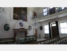 Sala storica con pareti bianche decorate da grandi dipinti e ritratti ovali, un grande camino in pietra e un lampadario di cristallo al centro. In alto corre una balconata con ringhiera in ferro battuto, mentre a destra si vedono finestre con tende e una porta in legno, con file di sedie rivolte verso un tavolo.