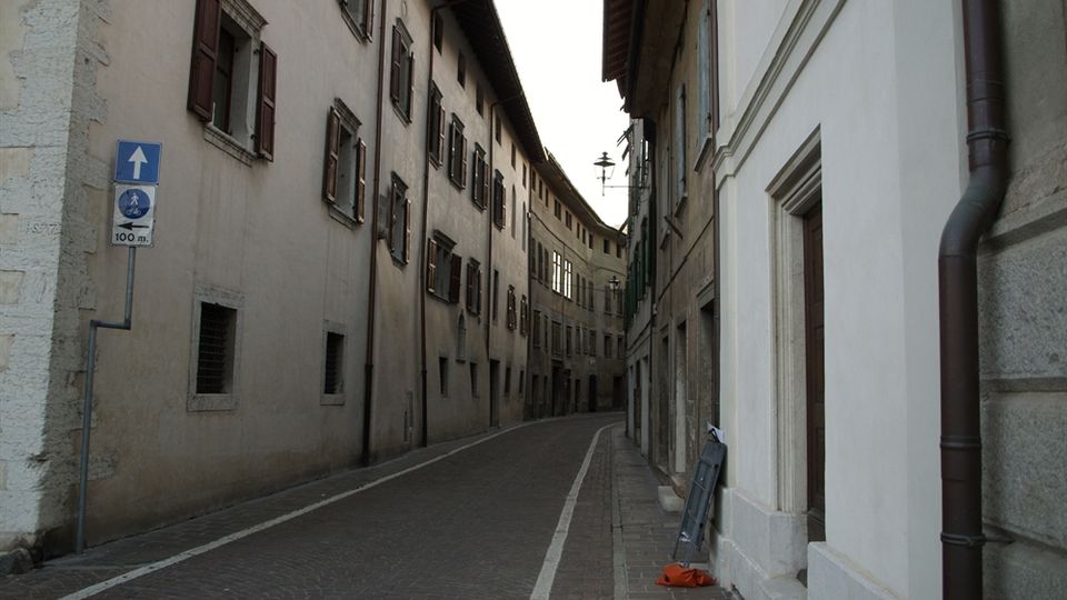 Strada stretta e curva in un centro storico, pavimentata in pietra e fiancheggiata da edifici alti con intonaco chiaro e finestre con scuri in legno. Sul lato destro si vede una facciata bianca con cornici in pietra e pluviale, mentre in fondo è presente un lampione a parete e segnaletica stradale.
