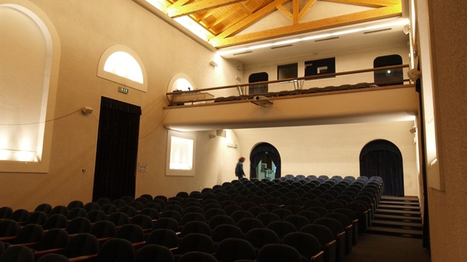 Interno di una piccola sala auditorium con file di poltrone scure, pareti chiare e finestre ad arco. Soffitto alto in legno con travi a vista e illuminazione scenica sospesa, con balconata laterale e palco sul fondo.