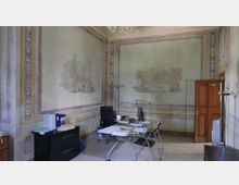 Ufficio in una stanza storica con pareti affrescate a riquadri e bordure decorative, con lampadario a soffitto e pavimento in graniglia. Al centro ci sono una scrivania in vetro con computer e documenti, sedie e mobili contenitori, con una porta in legno su un lato e una finestra con tenda sull’altro.