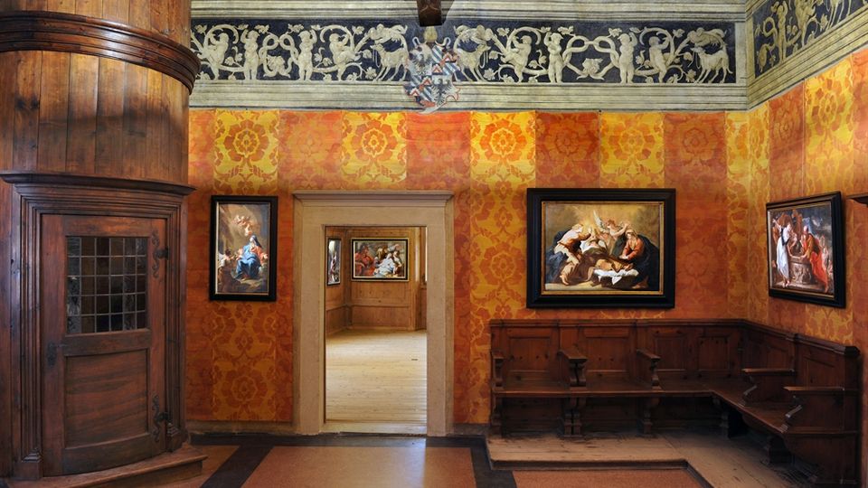 Interno di una sala storica con pareti rivestite in tessuto damascato arancione e una fascia decorativa superiore con motivi in rilievo, sotto un soffitto con travi in legno. Sulla sinistra si vede una grande stufa in legno, mentre lungo le pareti ci sono panche lignee e quadri incorniciati; al centro un portale conduce a un’altra stanza simile.