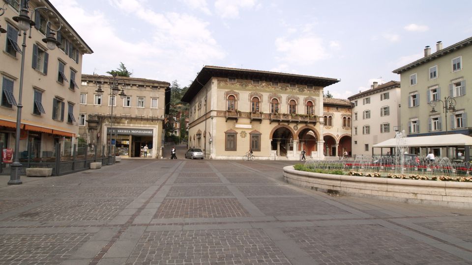 Ampia piazza lastricata in pietra, circondata da palazzi storici a più piani con persiane e lampioni. Al centro si vede un edificio decorato con portico ad archi e balconata, mentre sulla destra c’è una fontana circolare con aiuola fiorita e tavolini coperti da una struttura bianca.