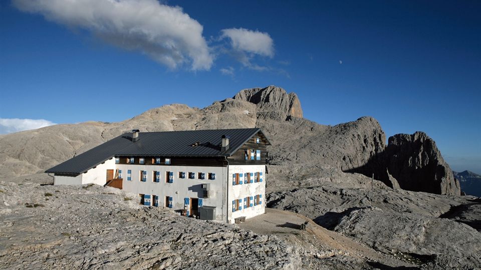 Rifugio di montagna in muratura con tetto scuro e file di finestre, situato su un altopiano roccioso. Sullo sfondo si vedono creste e pareti di montagne grigie sotto un cielo azzurro con poche nuvole.
