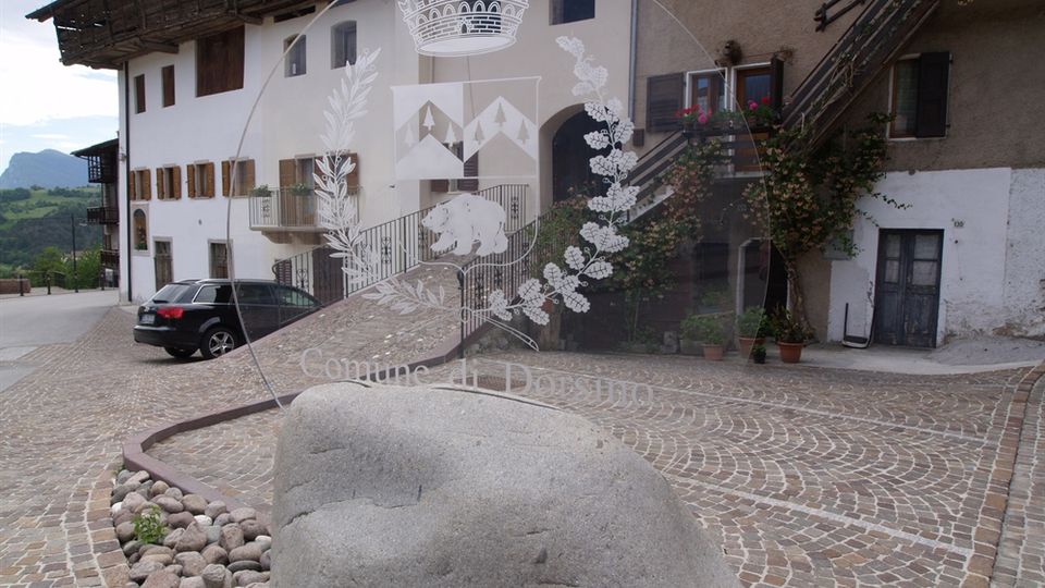 Piccola piazza lastricata in pietra con un grande masso arrotondato in primo piano e un’aiuola di ciottoli. Sullo sfondo si vedono edifici tradizionali con facciate intonacate, finestre con scuri in legno e una scala esterna in ferro, con colline e montagne all’orizzonte.