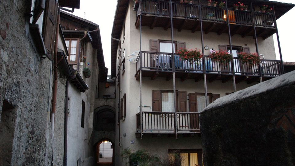 Vicolo stretto in un borgo storico, con case in intonaco e pietra affacciate su entrambi i lati. A destra un edificio con balconi in legno sovrapposti e fioriere, e in fondo un passaggio ad arco che conduce a una strada illuminata.