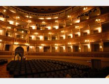 Interno di un teatro storico con platea di poltrone scure e più ordini di palchi curvilinei sovrapposti, decorati in legno dorato e illuminati da numerose luci calde. Sul lato sinistro si vede un ingresso con tendaggi e una porta ad arco.