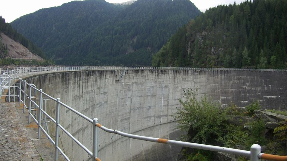 Diga in cemento ad arco vista dal camminamento con corrimano metallico e pavimentazione in pietra. Sullo sfondo si vedono montagne boscose e pendii rocciosi che circondano il bacino.